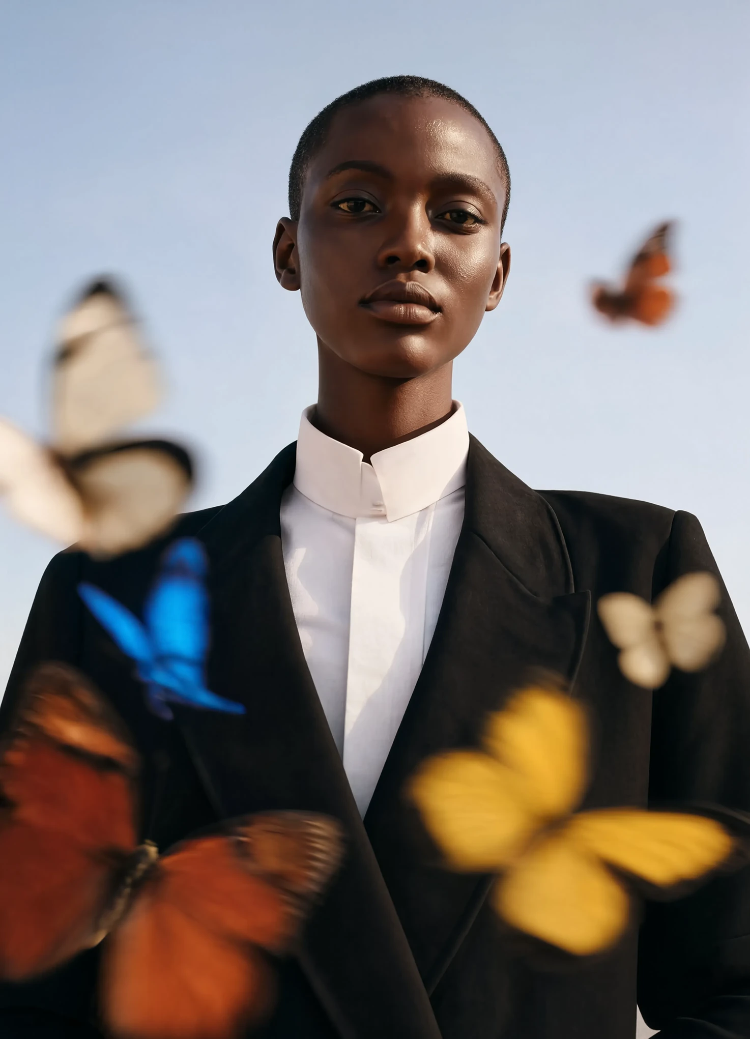 A woman in a black blazer and white shirt looks straight at the camera against a pale sky as butterflies blur past the lens
