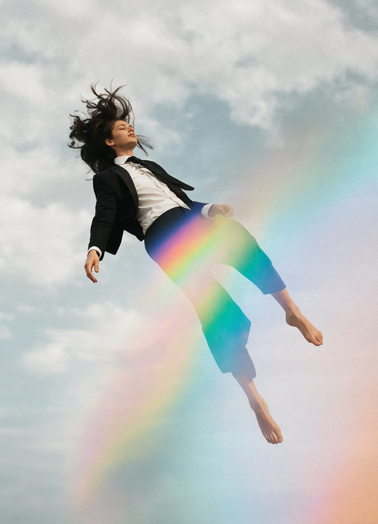 A woman in a black suit floating barefoot in mid-air among clouds, a rainbow crossing her legs