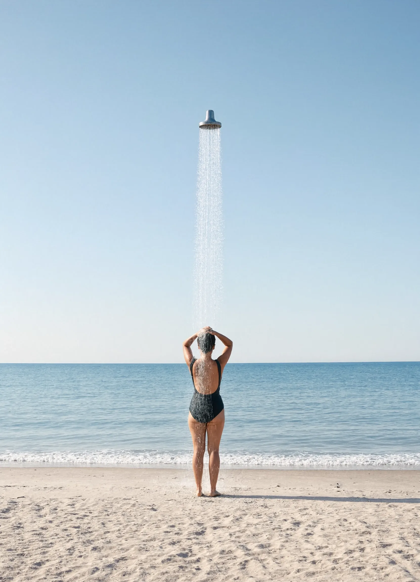 A woman stands on the shoreline beneath a shower head hanging unsupported in mid-air