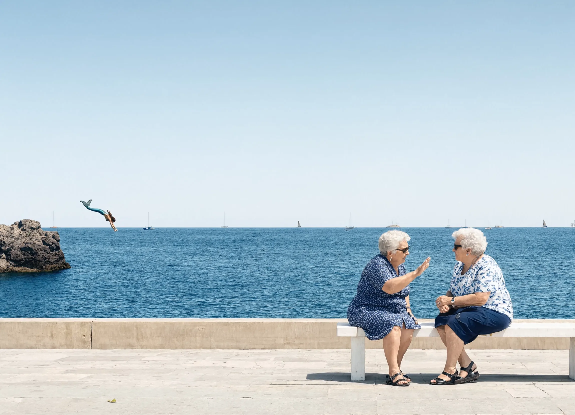 Two elderly women chat on a seafront bench while, unnoticed behind them, a mermaid dives from a rock into the sea