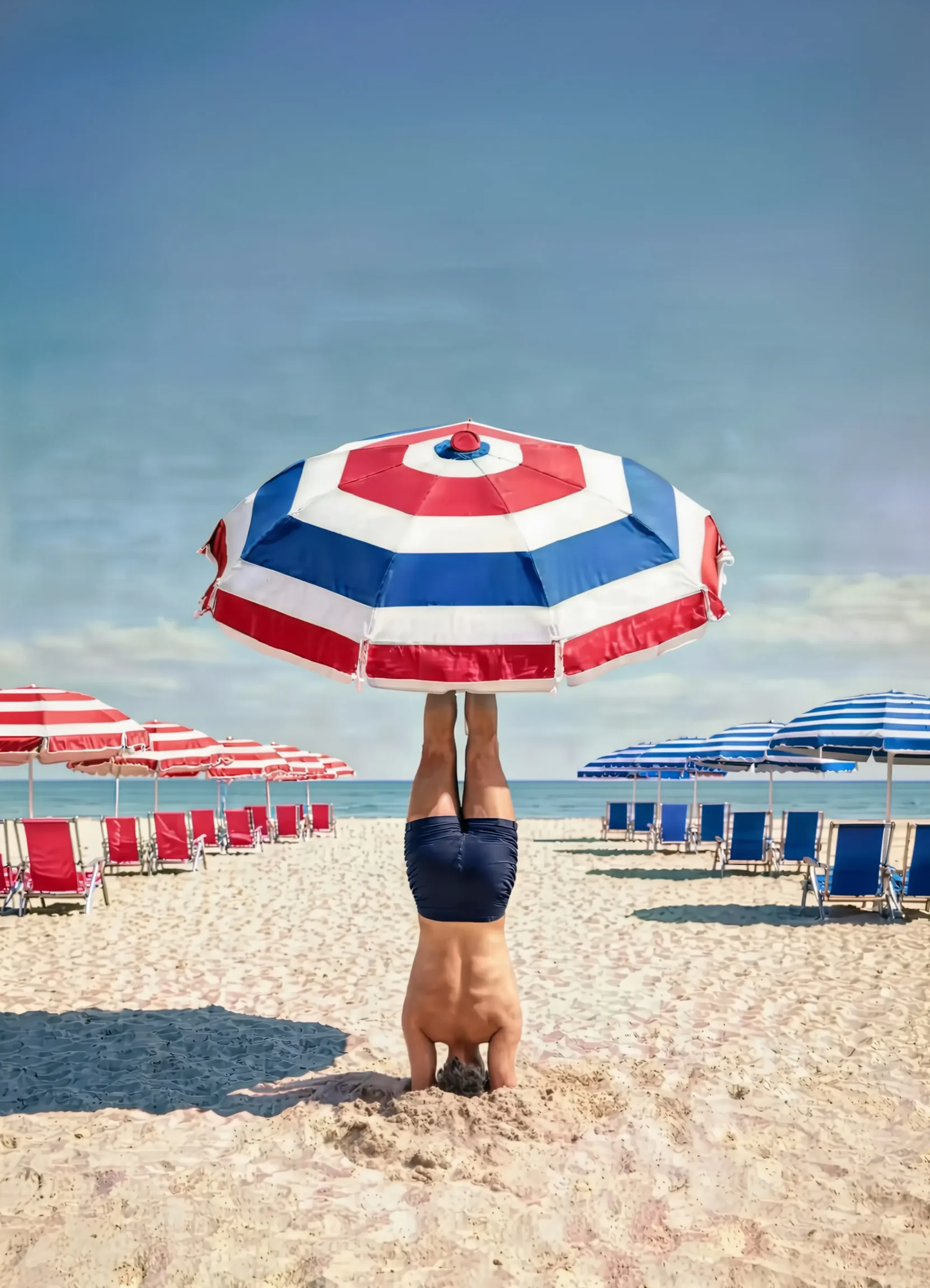 A man upside down with his head buried in the sand, balancing a red, white and blue beach umbrella on his feet, red loungers to the left and blue loungers to the right